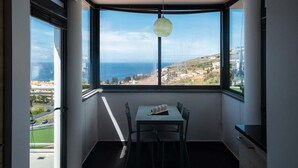 Ein Zimmer mit Tisch und Stühlen, einer Hängeleuchte und einem großen Fenster mit Blick auf das Meer und eine Stadt.