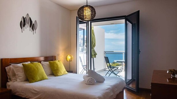Ein Schlafzimmer mit Bett, Kissen, einer Nachttischlampe und Meerblick durch eine offene Tür.