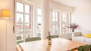 Ein heller Raum mit einem Holzesstisch, grünen Stühlen, einem weißen Sofa und großen Fenstern mit Blick auf Gebäude und eine Kuppelkonstruktion.