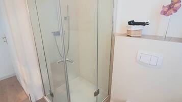 Ein modernes Badezimmer mit einer gläsernen Duschkabine, einer weißen Toilette und einer kleinen Ablage mit Korb und Pflanze.