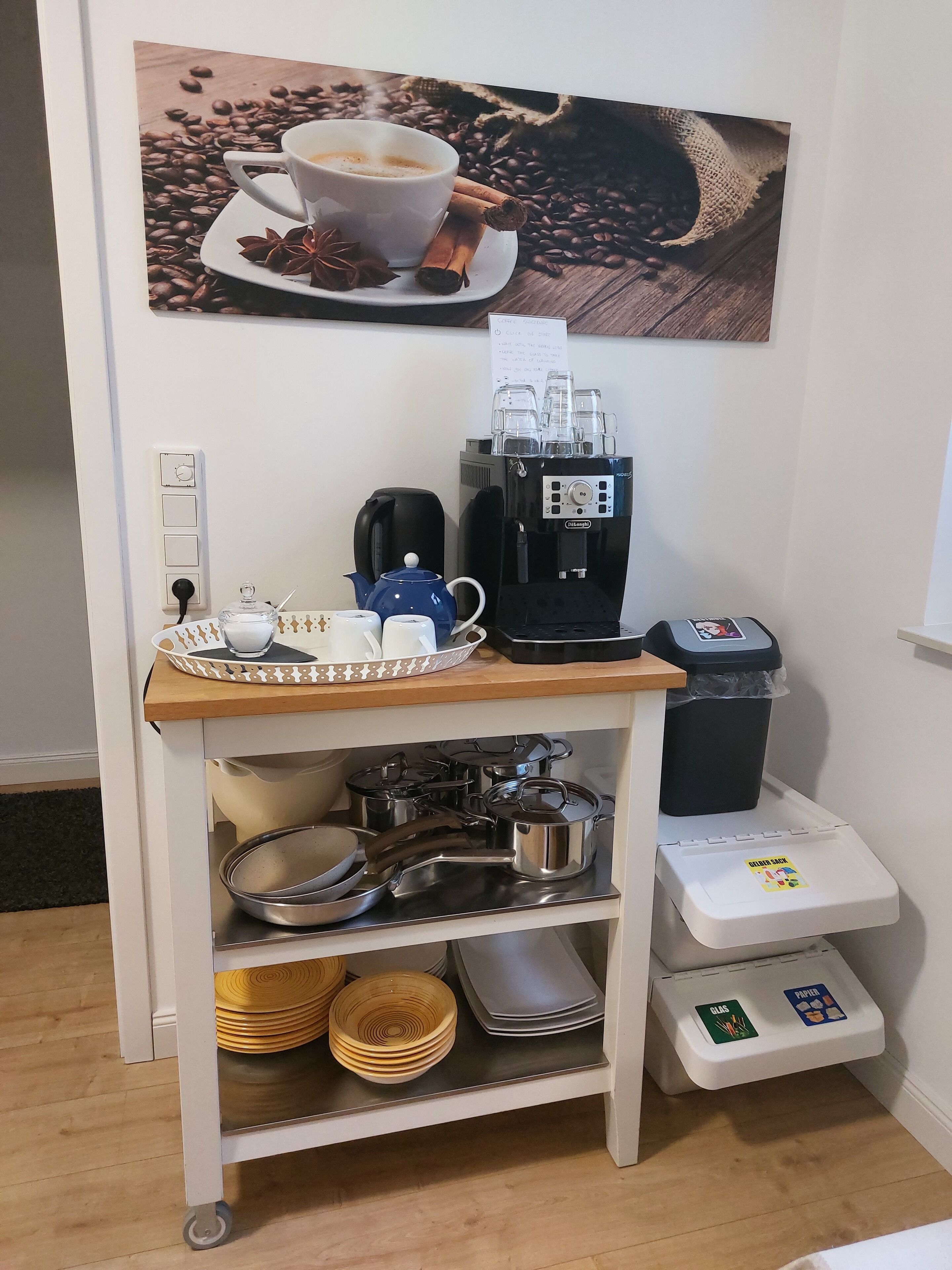 Uma cozinha compacta com cafeteira, chaleira, xícaras e lixeira.