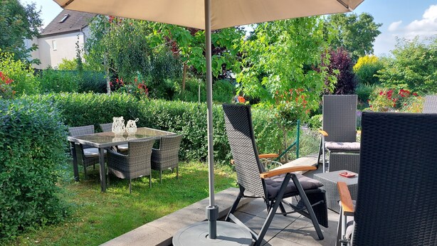 An outdoor patio area with a table and chairs, surrounded by lush greenery and a house in the background. - Moderne WHG in Churfranken: Fahrrad-wandertouren & Geschäftlich/monteuer😊 (Elsenfeld)
