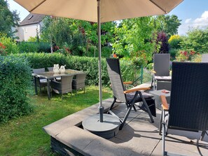 Un espace extérieur aménagé en terrasse, avec une table et des chaises, entouré d’une végétation luxuriante et avec une maison en arrière-plan.