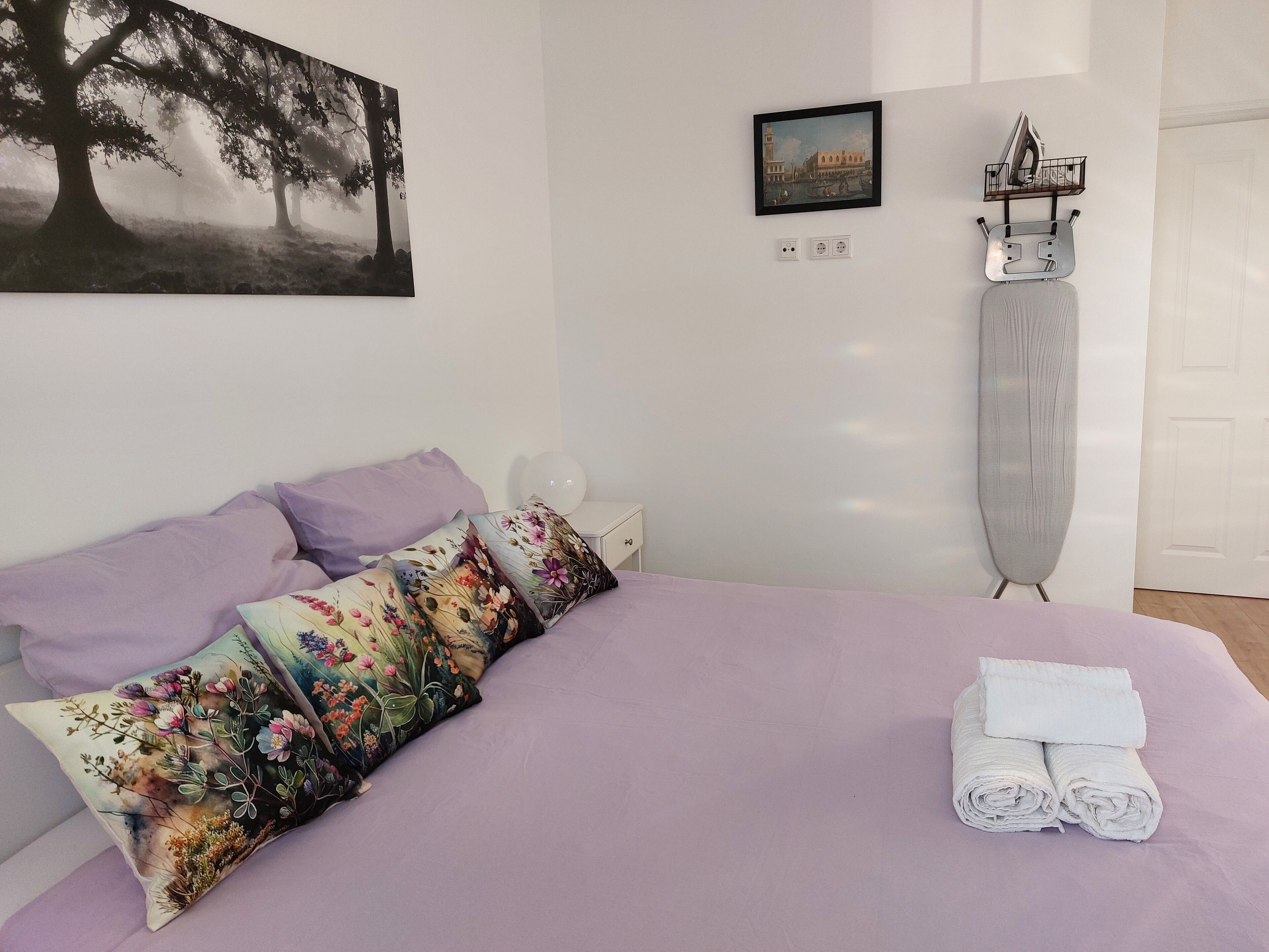 A bedroom with a bed, pillows, a painting, a small table, and a wall-mounted ironing board.