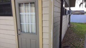 A beige single-story house with a gray front door, a small window with a white curtain, and a white fence in the background. - Cute, Quiet And Meticulously Kept. This Private Studio Is Close To Everything.
 (Brandon)