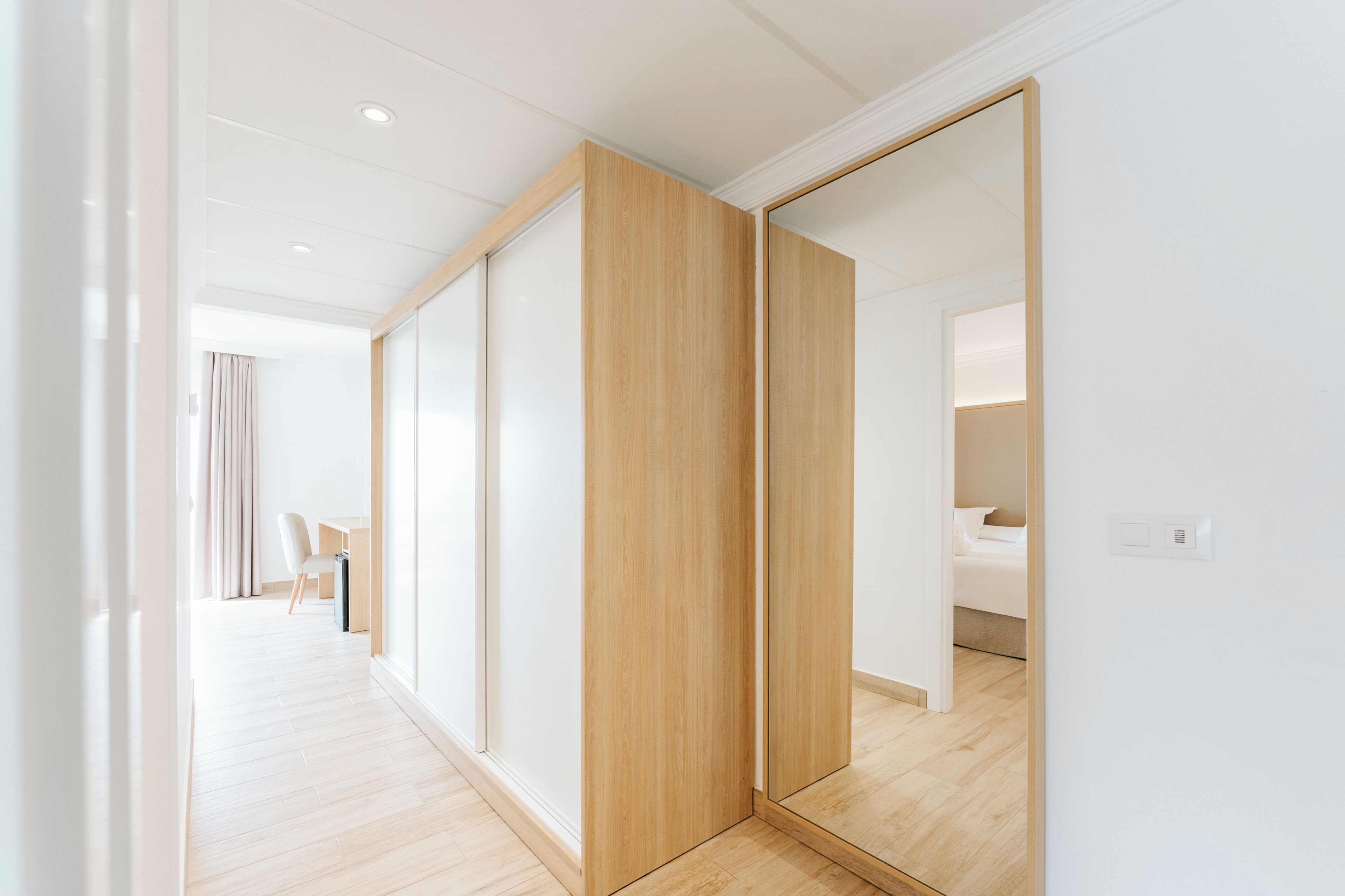 A modern hotel room with a large mirror, wooden flooring, and a view of the adjacent room.