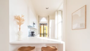 A modern kitchen with a white countertop, two wooden stools, a vase with dried plants, and a view of a yellow building through an open door. - GuestReady - Conde das Acáçovas 2E (Paço de Arcos)