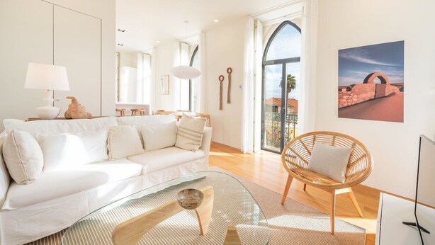A modern living room with a white sofa, a glass coffee table, and a wicker chair. There is a large window with a view of a building and a desert landscape. - GuestReady - Conde das Acáçovas 2E (Paço de Arcos)