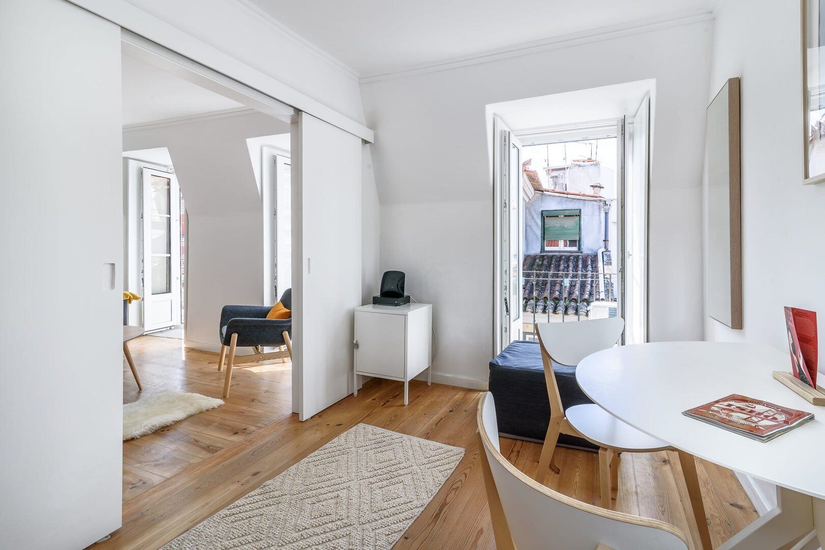 Un salón moderno con suelo de madera, un sofá blanco, una mesa de comedor con sillas y vistas a un balcón con barandilla.