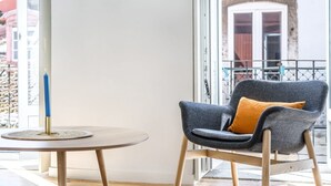 Une chambre moderne avec une table ronde en bois, une chaise rembourrée de couleur grise agrémentée d’un coussin moutarde, et une grande fenêtre donnant sur un balcon.