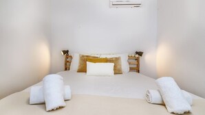 Un lit bien fait, avec du linge de lit blanc et deux serviettes enroulées, une table de chevet avec une lampe et un appareil de climatisation fixé au mur.