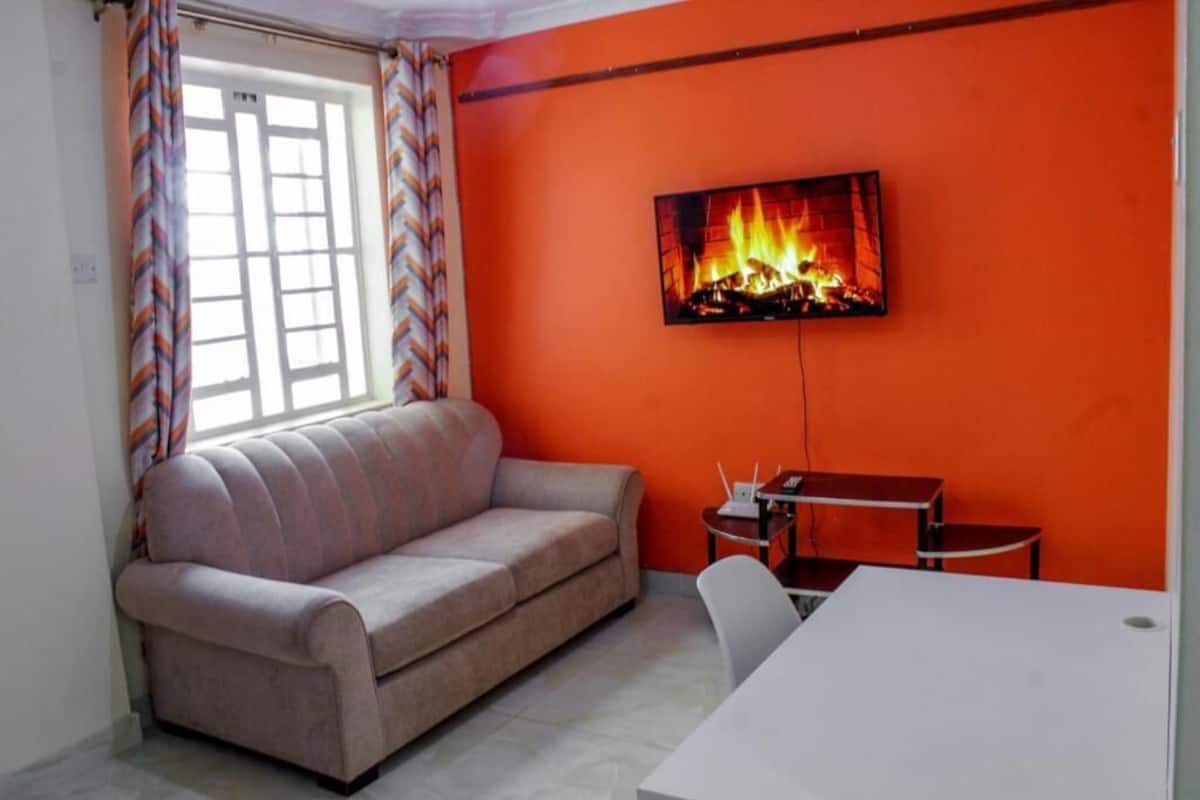 Un salón con sofá, televisor montado en una pared naranja, una mesita y una ventana con cortinas.