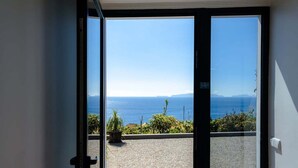 A view from a room with a glass door leading to a cobblestone patio overlooking a coastal landscape with a clear sky. - GuestReady - Leme Beach Breathtaking Views (Santa Cruz)