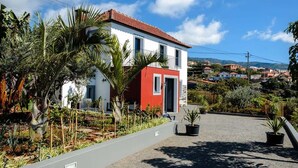 A two-story house with a red and white facade, surrounded by a garden with various plants and a paved driveway. - GuestReady - Leme Beach Breathtaking Views (Santa Cruz)