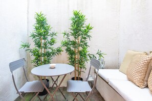 Un pequeño espacio al aire libre con una mesa redonda y dos sillas, un banco blanco acolchado y dos altas plantas de bambú.
