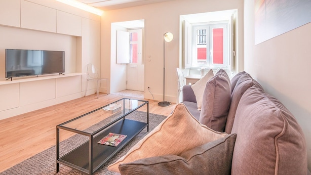 A modern living room with a flat-screen TV, a glass coffee table, a sofa, and a floor lamp. - GuestReady - Pink Sunset (Lisbon)