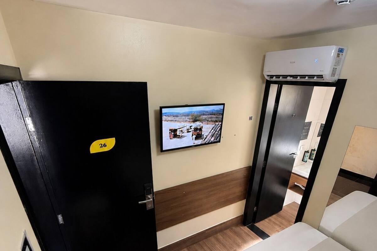 A hotel room with a bed, a television, a black door with a yellow sign, a white air conditioning unit, and a small white table.
