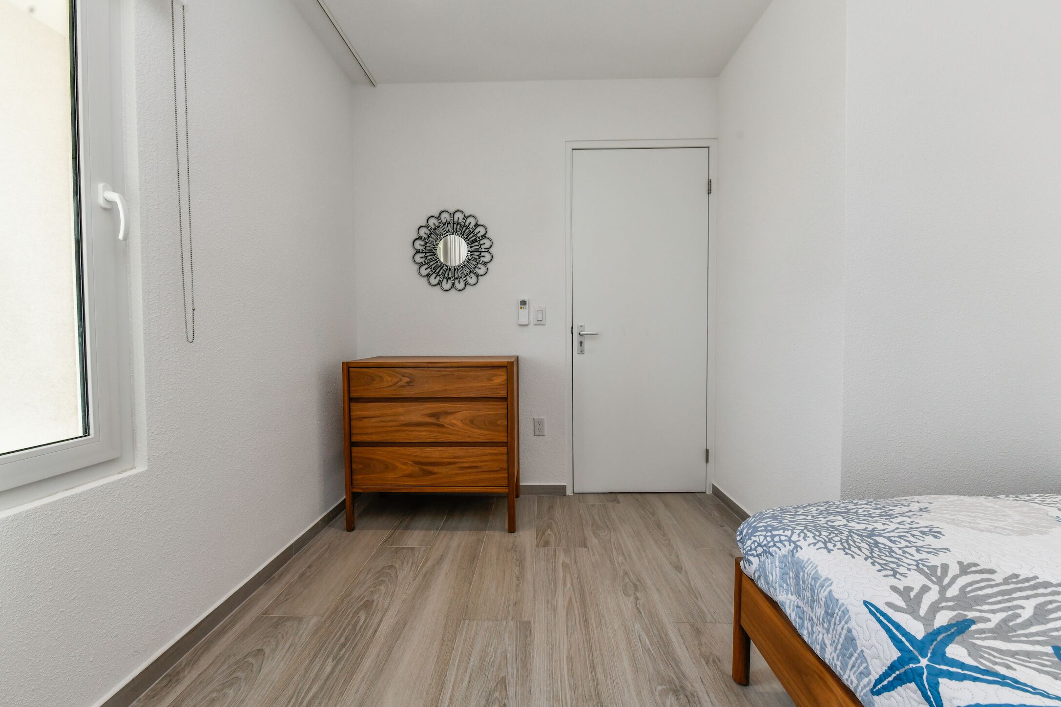 A bedroom with a wooden dresser, a bed with a starfish and coral pattern, a white door, and a wall-mounted mirror.