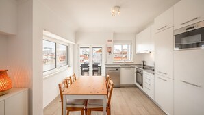 A modern kitchen with white cabinets, a wooden dining table, and a view of the city through large windows. - GuestReady - Sunny Retreat in Costa da Caparica (Costa da Caparica)