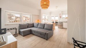 A modern apartment with a grey sectional sofa, a dining area, a kitchen, and large windows offering city views. - GuestReady - Sunny Retreat in Costa da Caparica (Costa da Caparica)