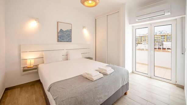 A hotel room with a bed, bedside lamps, a wall-mounted artwork, a balcony with a view, and an air conditioning unit. - GuestReady - Sunny Retreat in Costa da Caparica (Costa da Caparica)