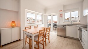 A modern kitchen with white cabinets, a wooden dining table, and a view of the city through large windows. - GuestReady - Sunny Retreat in Costa da Caparica (Costa da Caparica)