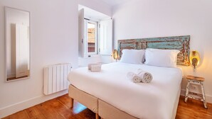 A bedroom with a large bed, a rustic headboard, a bedside table, a mirror, and a radiator. - GuestReady - Sunny Chic in Santos (Lisbon)