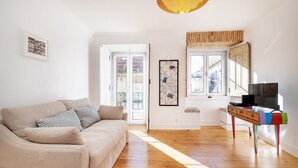 A living room with a beige sofa, a wooden floor, a TV on a colorful stand, and a large window with a view of buildings. - GuestReady - Sunny Chic in Santos (Lisbon)