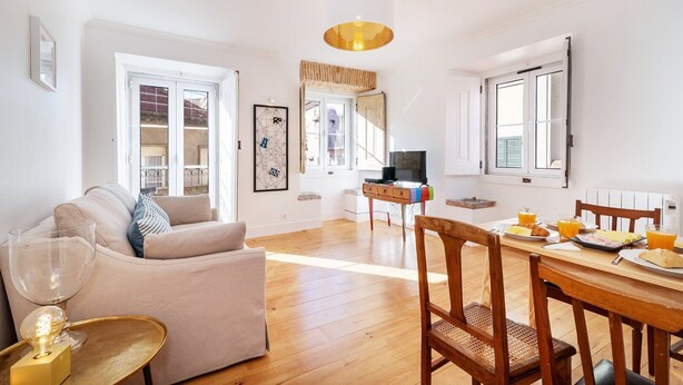 A spacious living area with a sofa, dining table, and chairs, a television, and a large window. - GuestReady - Sunny Chic in Santos (Lisbon)