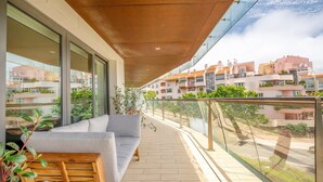 Un balcone con divano e sedie di legno, ampie finestre e vista sugli edifici circostanti.