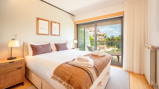 Camera d'albergo con un letto grande, un comodino, una lampada, una finestra con vista sugli edifici e un balcone con tavolo e sedie.