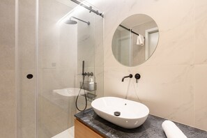 A modern bathroom with a glass-enclosed shower, a round mirror, and a white sink on a marble countertop.