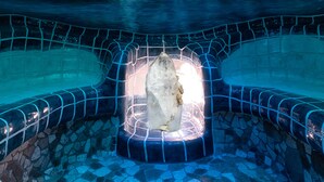 A hot tub with a stone sculpture in the center, surrounded by mosaic tiles, and a view of a river and mountains in the background.