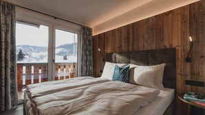 A bedroom with a large bed, wooden paneling on the walls, a view of snowy mountains through a window, and a small table with a lamp.