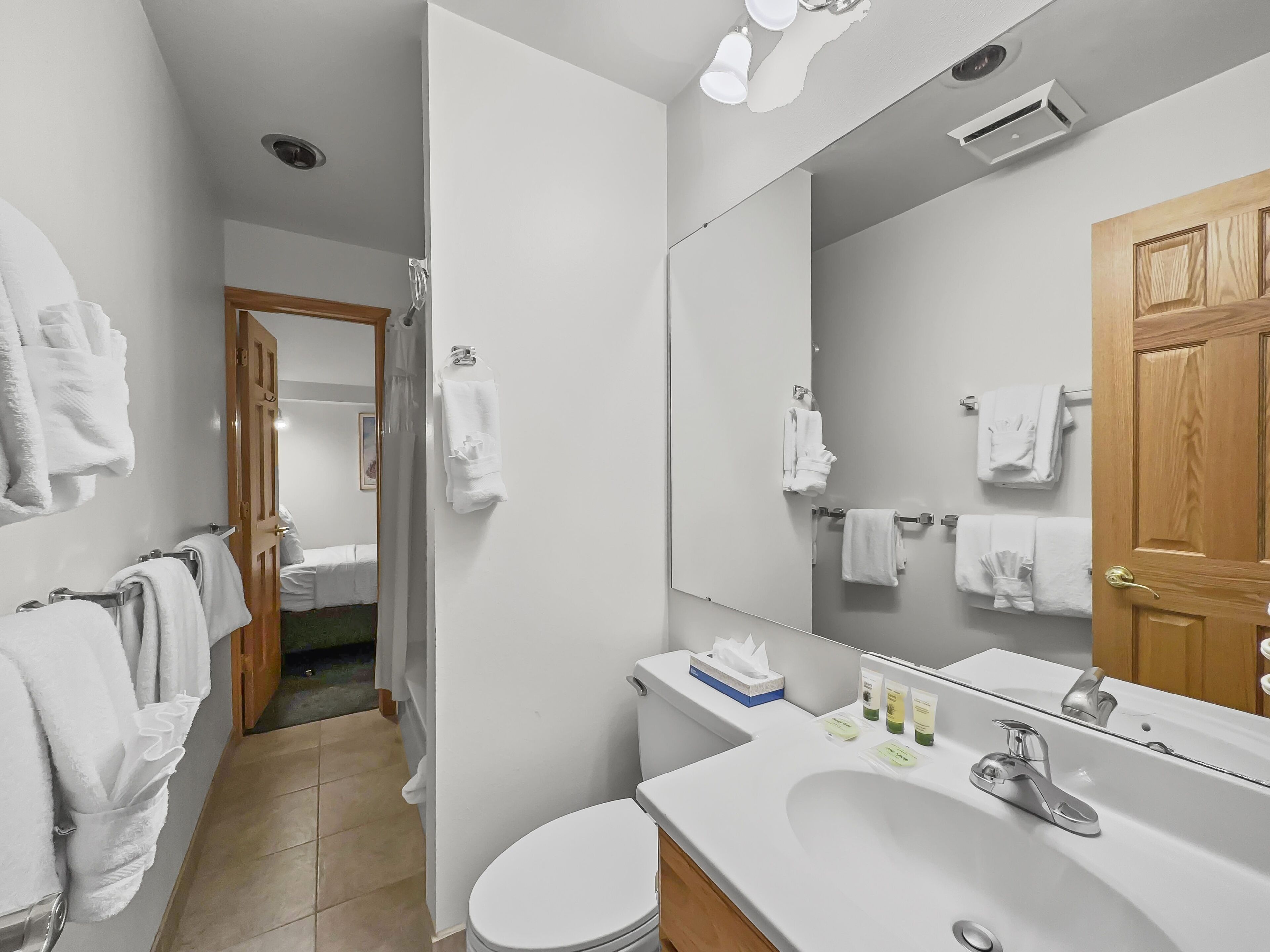 Une salle de bain moderne avec un lavabo blanc, un miroir et des toilettes. Plusieurs serviettes blanches sont accrochées au mur, et il y a une porte en bois.