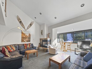 A modern living room with a fireplace, leather sofa, wooden coffee table, and a large window offering a view of trees and snow. - Cj404 Copper Junction 3 Bedroom Condo by RedAwning (Copper Mountain)