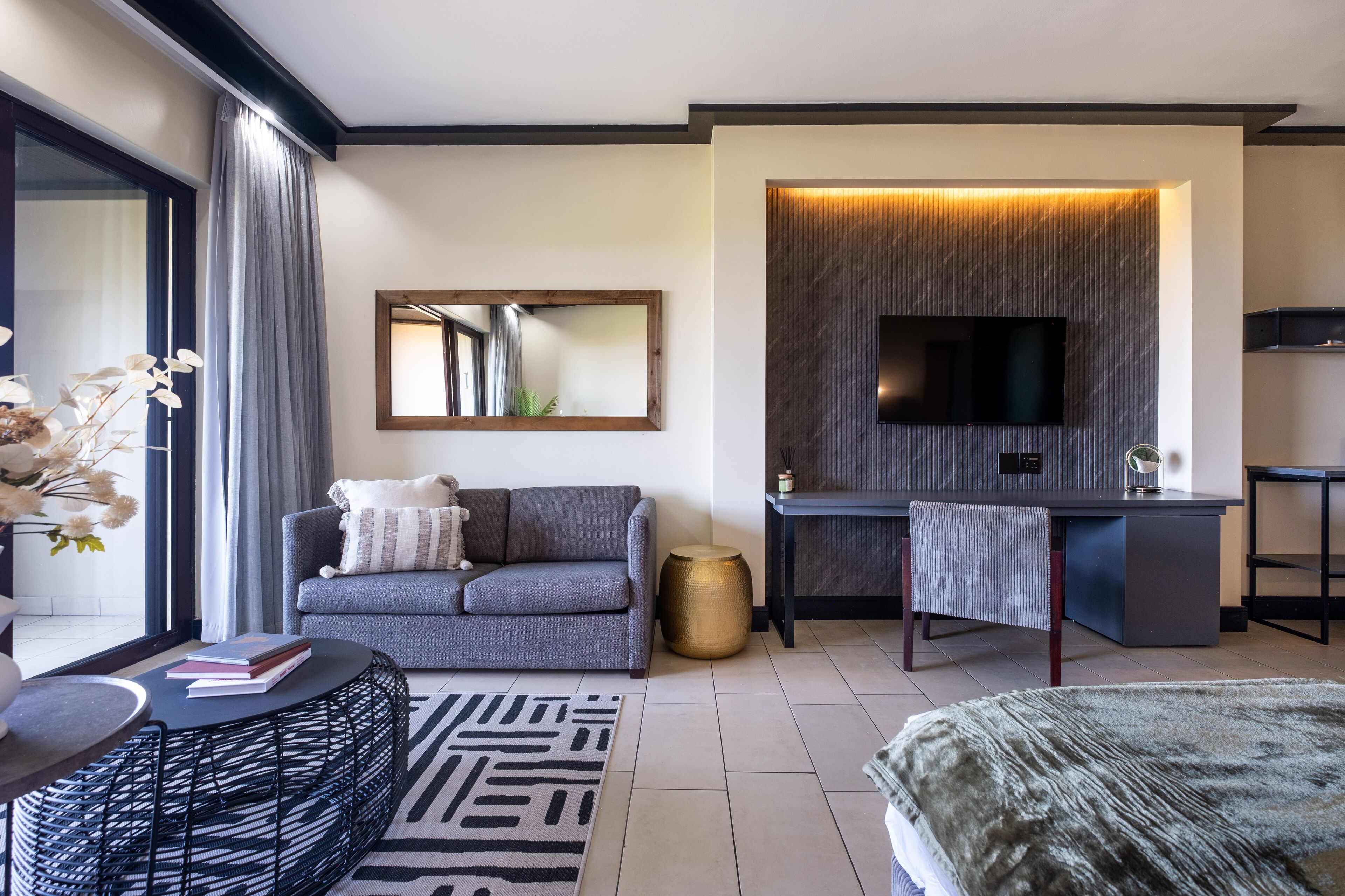 A modern hotel room with a grey sofa, a round black table, a TV mounted on a textured wall, and a small round side table.