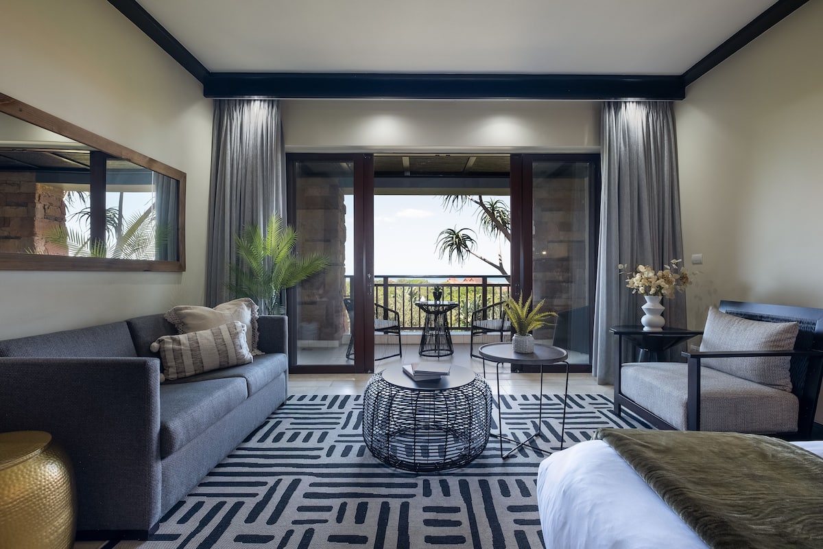 A modern hotel room with a sofa, a round coffee table, a bed, and a balcony with a view of palm trees.
