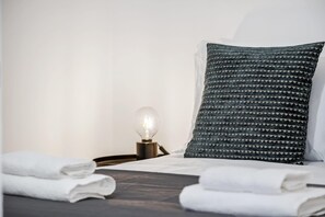 Una cama bien hecha con toallas blancas dobladas, un cabecero de madera oscura y una lámpara de noche con pantalla de cristal transparente.