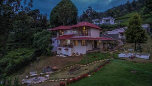 A two-story house with a red roof, surrounded by lush greenery and a garden with a pathway leading to the entrance. - Xplore Indo - Onella (Kotagiri)