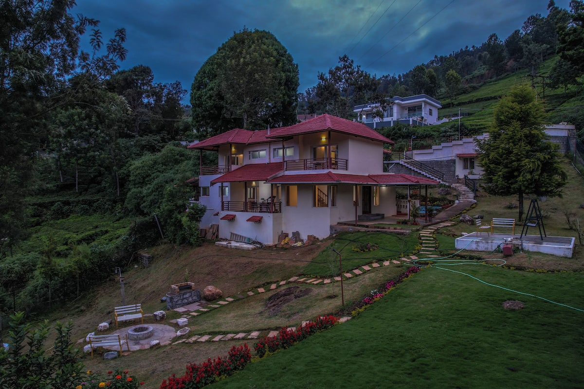 A two-story house with a red roof, surrounded by lush greenery and a garden with a pathway leading to the entrance.