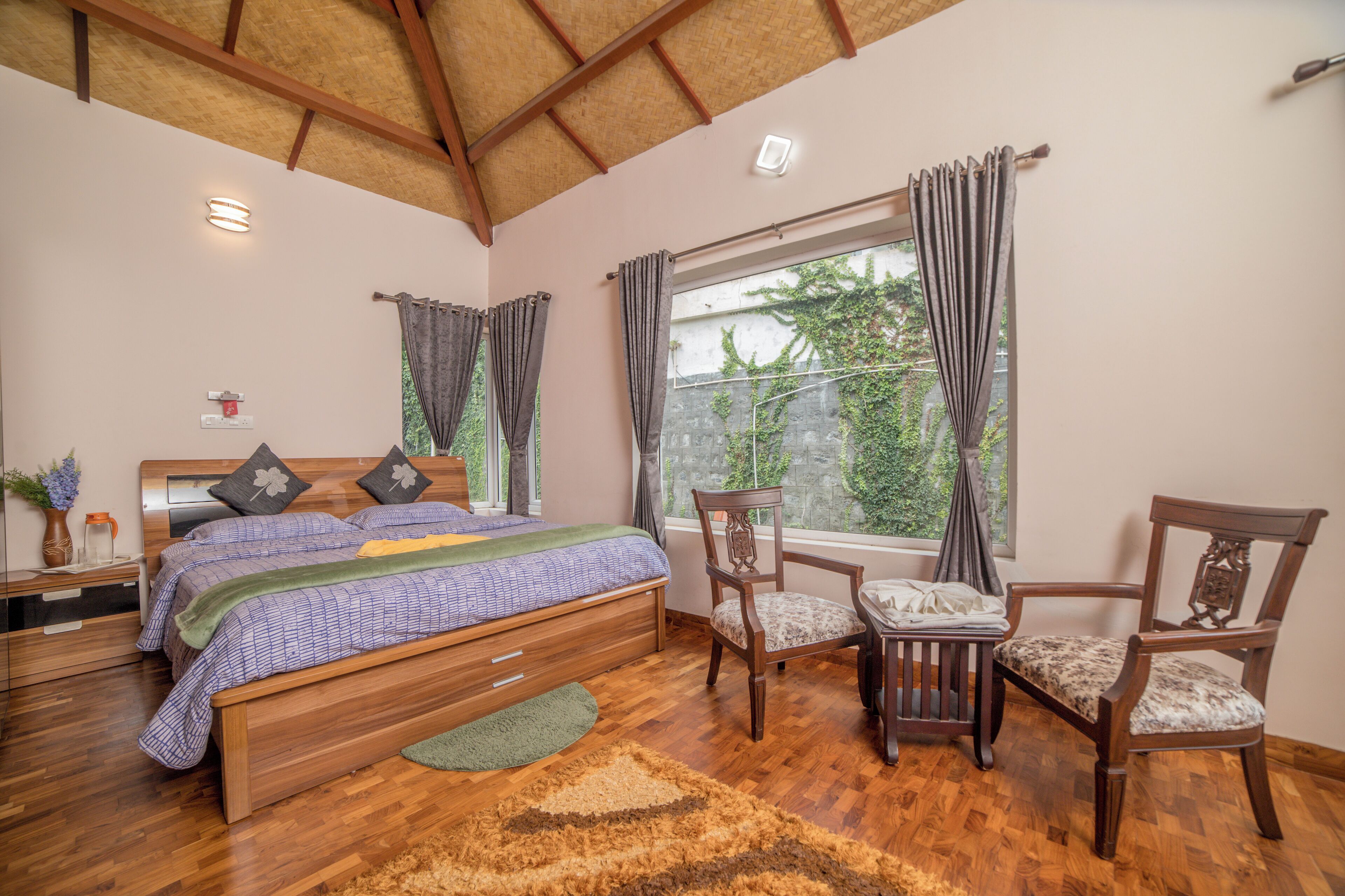 A bedroom with a wooden bed, two chairs, a window with curtains, a bedside table, and a nightstand.