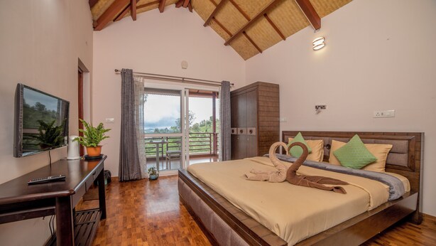 A bedroom with a wooden bed, a desk, a television, and a balcony with a view of the outdoors. - Xplore Indo - Onella (Kotagiri)