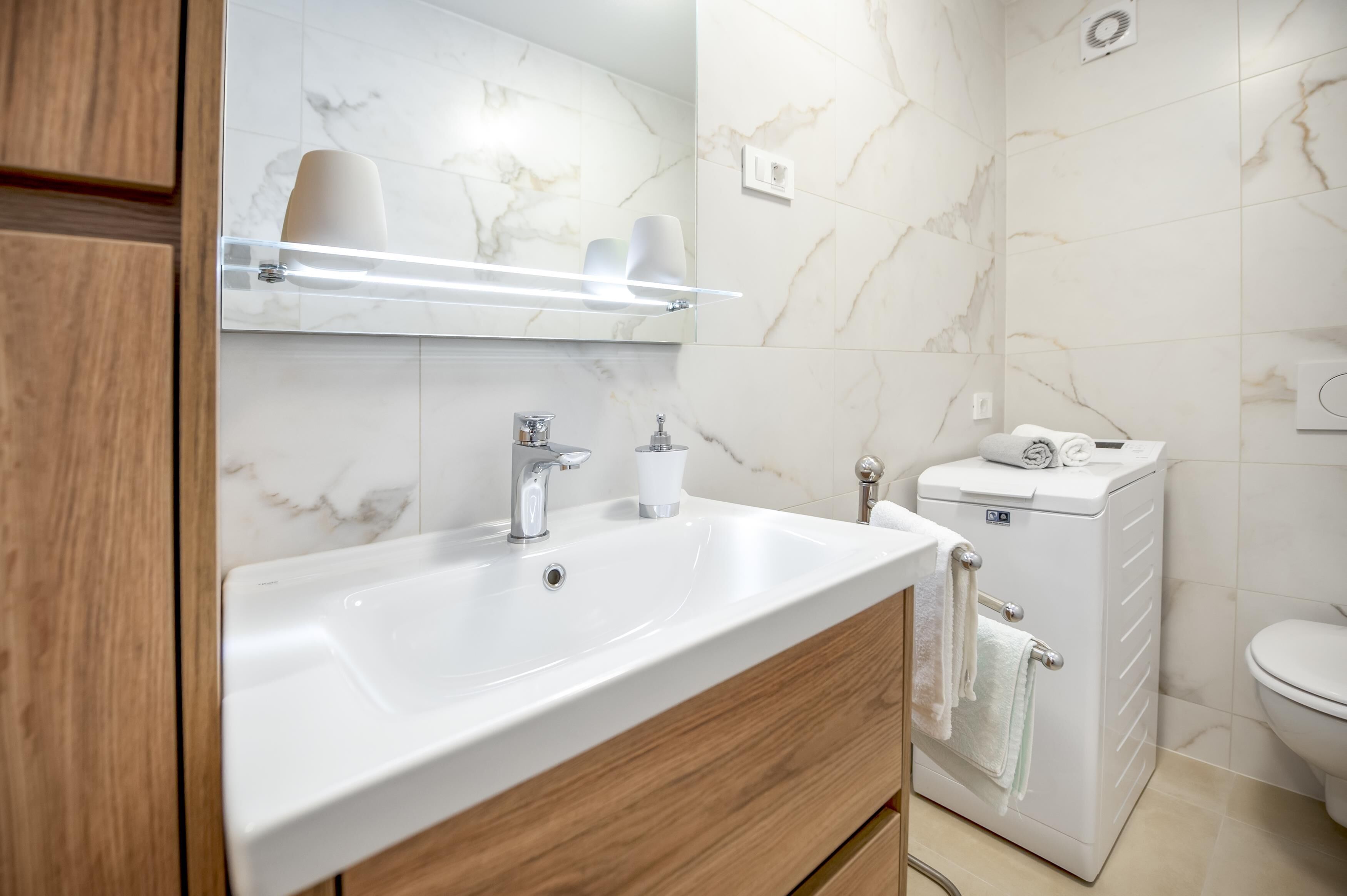 Un bagno moderno con un ampio lavandino bianco, mobili in legno, uno specchio con illuminazione a LED, un water e un porta asciugamani.