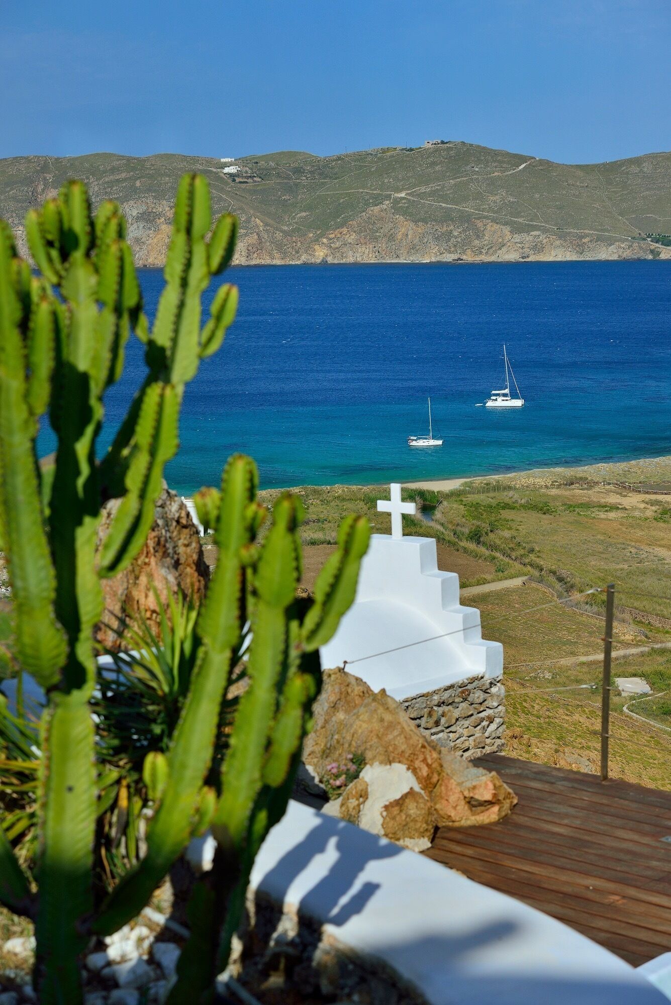 A coastal view with a white church, a cactus in the foreground, and a sailboat on the sea.