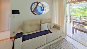 A modern living room with a beige sofa, a round wall clock, and a view of greenery through large windows. - Paraíso na Reserva Imbassaí (Mata de São João)