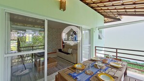 A dining area with a wooden table set for four, a sofa, and a view of greenery outside. - Paraíso na Reserva Imbassaí (Mata de São João)