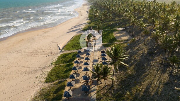 An aerial view of a beach resort with a long pathway leading to a building, surrounded by palm trees and beach umbrellas. - Paraíso na Reserva Imbassaí (Mata de São João)