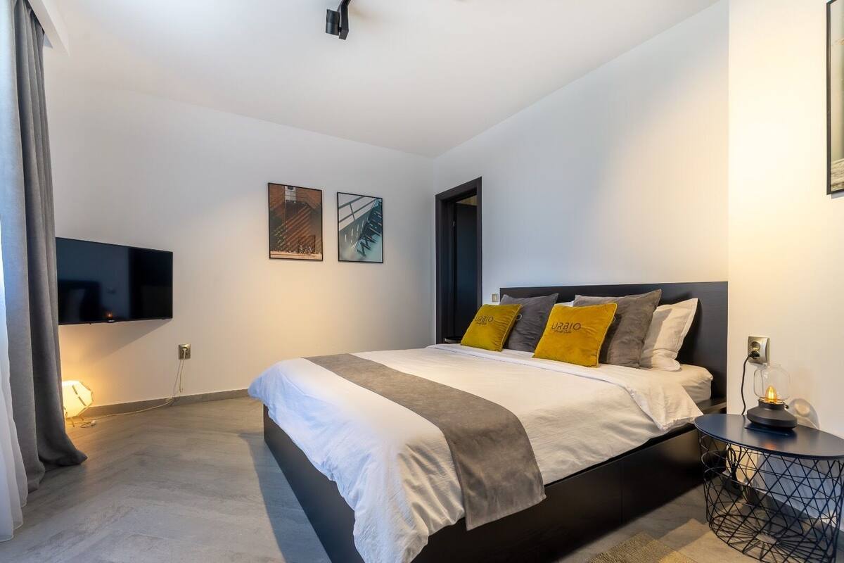 A modern bedroom with a large bed, a flat-screen TV mounted on the wall, and a bedside table with a lamp.