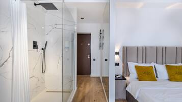 A modern hotel room with a glass shower enclosure, a wooden floor, a bed with two pillows, and a nightstand with a lamp.